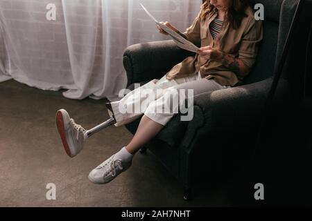 Beautiful young woman with prosthetic leg reading book on sofa in