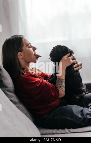 Side view of young woman sticking out tongue to pug dog on sofa Stock Photo
