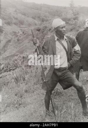 Kenya, A young man in a tattered jacket stands barefoot on a hillside, holding a spear. A white cloth around his arm and head indicates he is a member of the Kikuyu Home Guard. Original typescript caption: A Kikuyu Home Guard on duty, during the round-up of a gang which had come out of the forest. The Guards tie bits of cloth round their heads to distinguish friend from foe. Many of them are armed with spears and swords, a few with rifles. The[y] are volunteers and unpaid and have killed or captured many hundreds of armed gangsters, 1953. 1995/076/7/8/2.11. Stock Photo