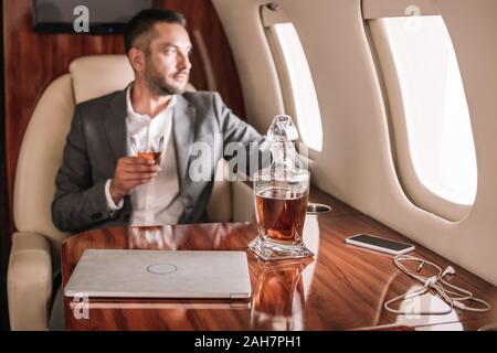 selective focus of gadgets and jar with whisky near bearded businessman in private jet Stock Photo