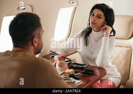 selective focus of woman sitting with man holding chopsticks near sushi in private jet Stock Photo