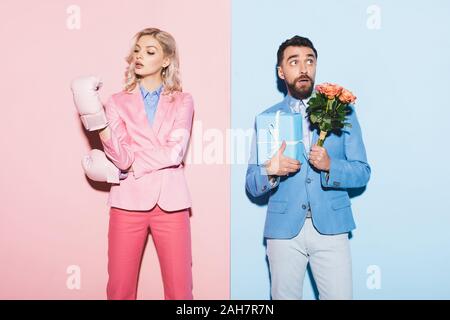 serious woman with boxing gloves and scared man with gift and bouquet on pink and blue background Stock Photo