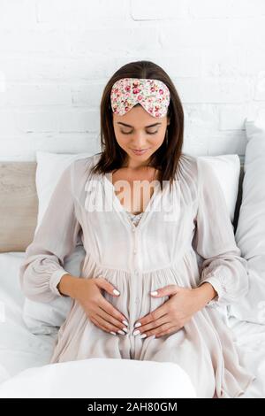 pregnant woman in sleeping mask touching her belly while lying in bed Stock Photo