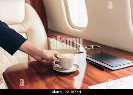 cropped view of flight attendant putting cup of coffee on table in private plane Stock Photo