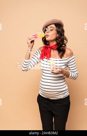 pregnant french woman in beret blowing soap bubbles on beige background Stock Photo