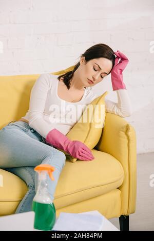 tired housewife sitting on yellow sofa with closed eyes near table with spray bottle Stock Photo