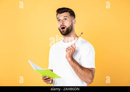 shocked handsome man in white t-shirt with open mouth holding pencil and notebook isolated on yellow Stock Photo