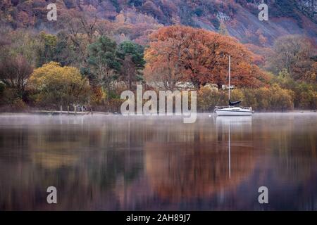 Moored yacht and reflections with mist rising on Derwent Water at Autumn time in the English Lake District, Cumbria. Stock Photo