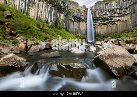 Low angle long exposure of the iconic Icelandic waterfall of Svartifoss, with its water flowing between pebbles and moss-covered rocks Stock Photo