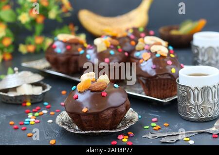 Banana-chocolate muffins, covered with icing and confectionery confetti are located on a dark background Stock Photo