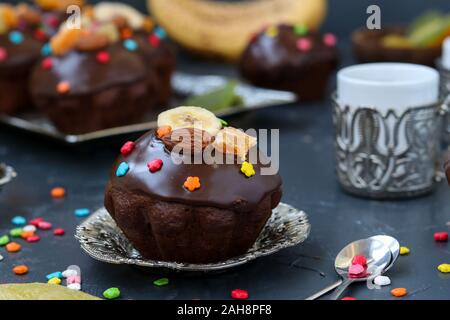 Banana-chocolate muffins, covered with icing and confectionery confetti, are located on a dark background Stock Photo