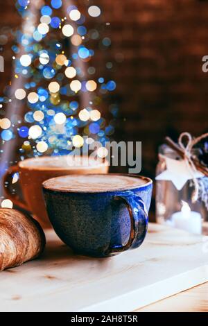 Perfect breakfast of croissants and cappuccino in a cafe. Blue and yellow ceramic cups of cappuccino and croissants on wooden table with Christmas lig Stock Photo
