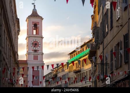 Evening view of the Rusca Palace Clock Tower at the Place du Palais du Justice with red and white flags draped over the street in Nice, France Stock Photo