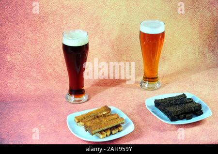 Two tall glasses with light and dark beer stand in front of two plates of fresh wheat and rye croutons on a brown background. Close-up. Stock Photo