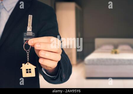 Man shows keys with keychain in form of a little house into a new flat. blurred background of the bedroom room with a modern interior. Stock Photo