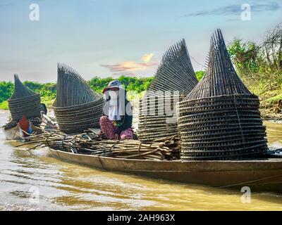 A Cambodian woman sits in a wooden boat being towed down a river. The boat is filled with homemade fishing trap. Stock Photo