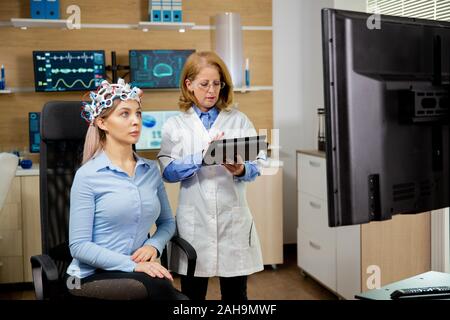 Patient woman scanning her brain and doctor making notes in the tablet holding it in her hand. Brain waves scanning device Stock Photo