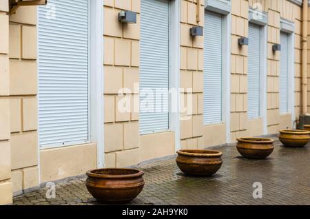 Five shop windows are closed by shutters. Four empty ceramic flowerpots for street flowers near the store. Wet sidewalk after the rain. Without people Stock Photo