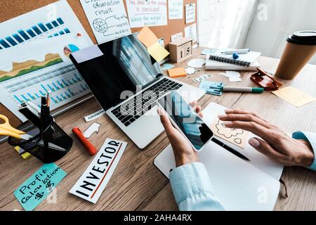 cropped view of businesswoman holding smartphone with blank screen near notebook and laptop in office Stock Photo