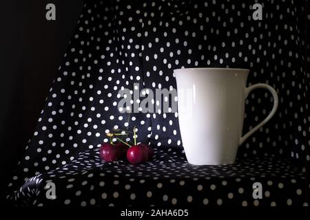 red cherry with white ceramic mug for hot drink on polka white dot on black fabric dark still life background Stock Photo
