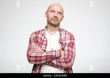 Frowning bald man stands with his arms crossed and looks thoughtfully upstairs, as if looking for a solution to the problem. Stock Photo