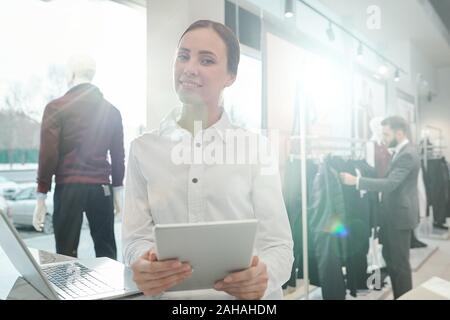 Pretty cheerful shop assistant in white shirt using touchpad by workplace Stock Photo