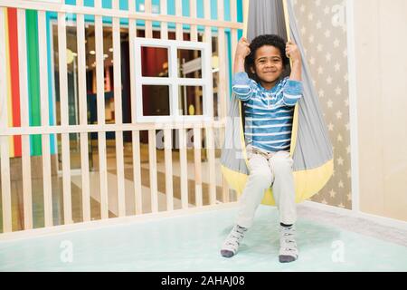 Happy little African boy in casualwear sitting in hammock hanging from ceiling Stock Photo