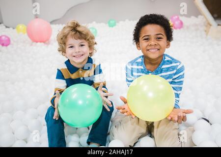 Cute cheerful intercultural little boys in striped shirts playing with balloons Stock Photo