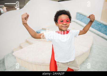 Strong cute little boy of African ethnicity wearing red mantle of super hero Stock Photo