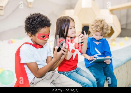 Cute African boy in costume of superhero and his two friends using gadgets Stock Photo