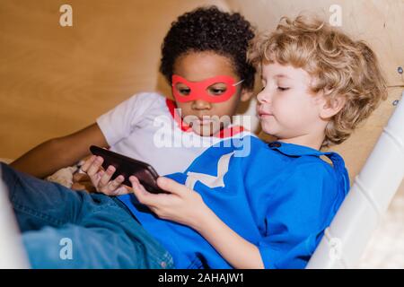 Two boys in costumes of superman looking at screen of smartphone while relaxing Stock Photo