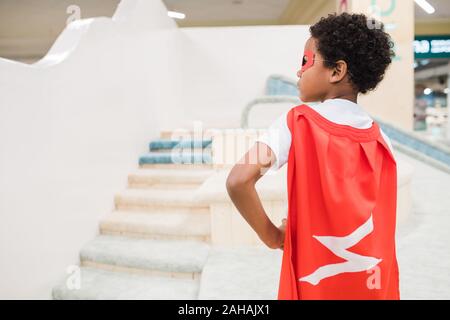 Back view of little African boy in costume of superman standing on play area