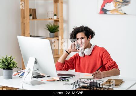 Young smiling freelancer sitting in front of computer and talking on smartphone Stock Photo