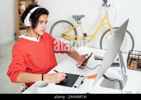 Contemporary guy with headphones using smartphone in front of computer Stock Photo