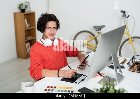 Happy guy with stylus and smartphone looking at you while using graphics tablet Stock Photo