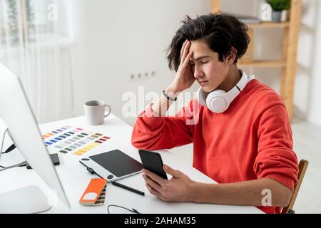 Tired freelancer touching his head while using smartphone in front of computer Stock Photo