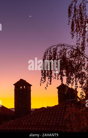 Waning crescent moon, dawn purple and orange colorful background with tree and houses silhouette Stock Photo