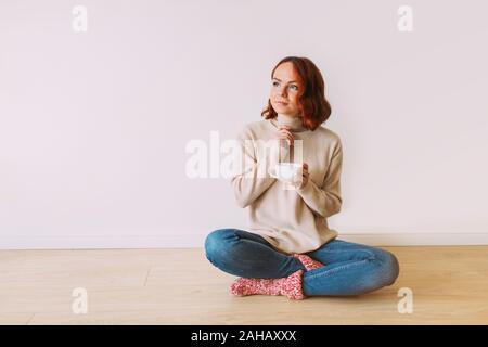 Dreaming young red haired girl sitting on the floor on a white blank background. Female model looking up, holding a mug with hot coffee. Copy space. Stock Photo