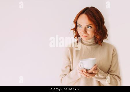 Lifestyle image of red haired beautiful girl sipping coffee from a white mug, wearing cozy sweater of a pastel color. Looking aside. Copy space. Stock Photo