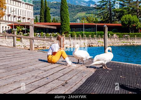 Riva del Garda, Lombardy, Italy - September 12, 2019: Tourist girl sitting on a wooden pier with two white swans walking around on promenade of Garda