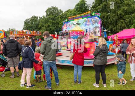 People watching the Cliffhanger fairground ride and a children's tea cups ride in wet weather in summer after the Thelwall Rose Queen procession 2019 Stock Photo