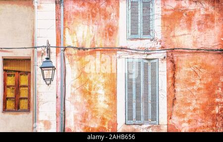 An old, cracked building wall with peeling paint and grunge textures, closed wooden shutters, and an electric hanging lantern. Provence, France. Stock Photo