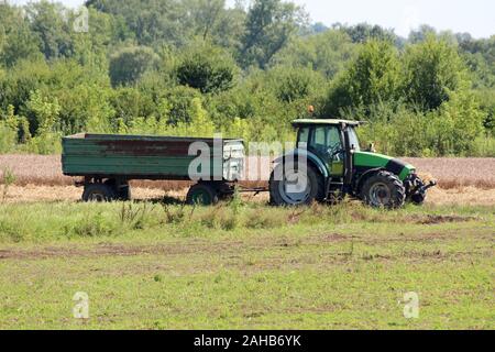 Green and black farm tractor with connected rusted vintage old tractor trailer left in local field surrounded with uncut grass and ripe wheat Stock Photo
