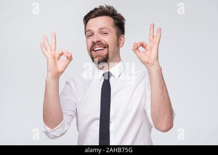 Happy young man in shirt and tie gesturing approving sign and smiling. Stock Photo