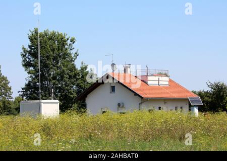 Weather station building with new roof tiles surrounded with various weather instruments and tall grass filled with small flowers on clear blue sky Stock Photo