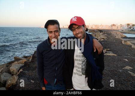 Two young Tunisian men standing by the seashore near the whitewashed buildings of the old city of Mahdia on the Mediterranean coast of Tunisia. Stock Photo