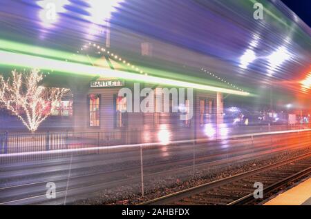 Bartlett, Illinois, USA. Railroad station decorated for Christmas is visible through the lights and steel of a passing train providing an almost dream Stock Photo