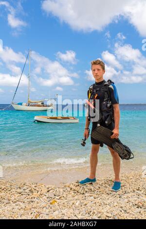 European male diver standing on beach of Bonaire with sea and boats Stock Photo
