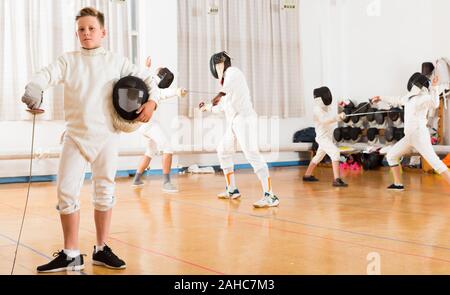 Portrait of nice boy wearing fencing uniform standing in gym with foil and mask in hands Stock Photo