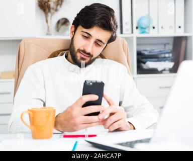 Young positive office worker using his smartphone at workplace in office Stock Photo